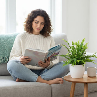 Woman reading a book on a couch with a Boston Fern and cup on a table beside her